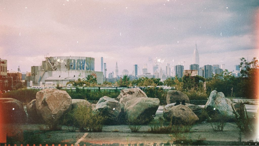 A vintage colour photograph of an urban cityscape with large stones in the foreground, gas storage facility covered in graffiti behind to the left and a city skyline in the distance. The photograph has a large number of scratches, light leaks and other imperfections along with faded colour.