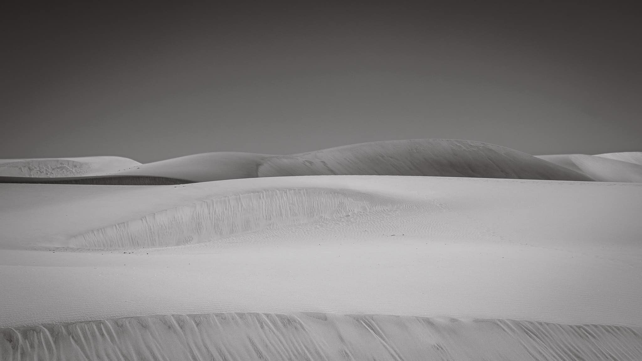 Black and white photo of Grand Canaria desert landscape featuring several sand dunes with smooth, flowing lines. The dunes create a pattern of light and shadow with visible ripples and waves formed by the wind.