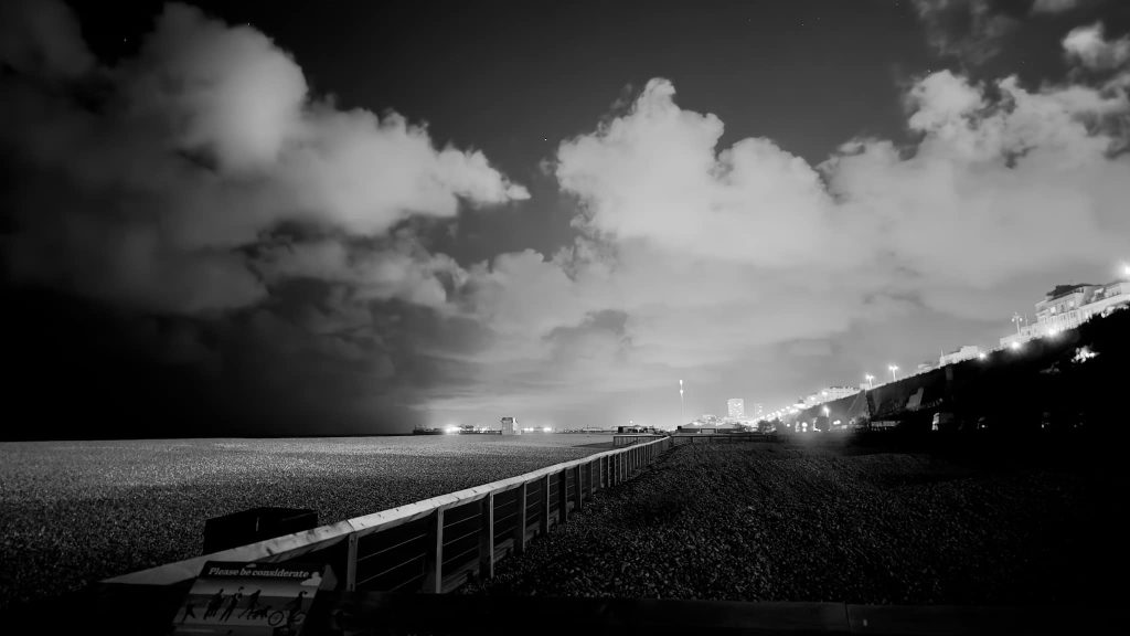 A black and white photo of Brighton beach at night with a fence in the foreground, city lights in the distance and a dark sky above.